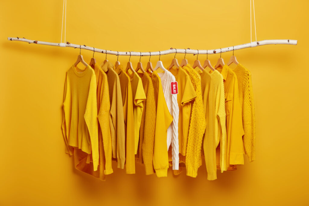 Black Friday, shopping and big sales concept. Detail image of yellow clothes and white sweater with label tag hanging on rack in clothing store. Shopping mall suggests customers season discounts